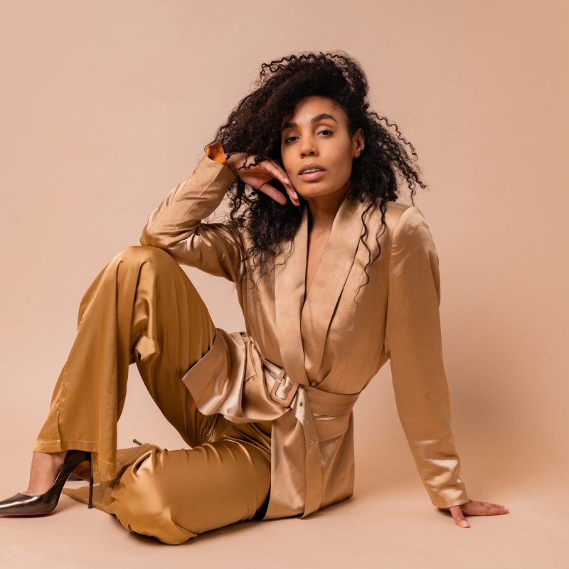 Gorgeous  black woman with beautiful wavy hairs in elegant  golden satin suit posing over beige background. Spring fashion look.